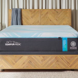 A well-lit bedroom features a wooden bed frame with a chevron pattern. The bed is made with a Tempur-Pedic TEMPUR-LuxeBreeze® Medium Hybrid 13" Mattress that has a black and blue design. The room has light-colored curtains, a gray cover draped over the headboard, and a small bedside table with decor.