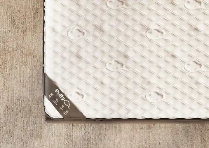 A corner of a white quilted Puffy LUX Mattress placed on a beige floor, with a label visible indicating its brand and qualities.