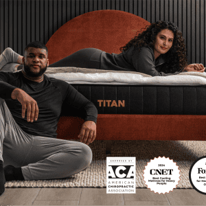 In a contemporary bedroom, a man sits on the floor while a woman relaxes on the Titan Plus mattress, showcasing its features. The display also highlights three awards from ACA, CNET, and Forbes for the Titan Plus's comfort and support.