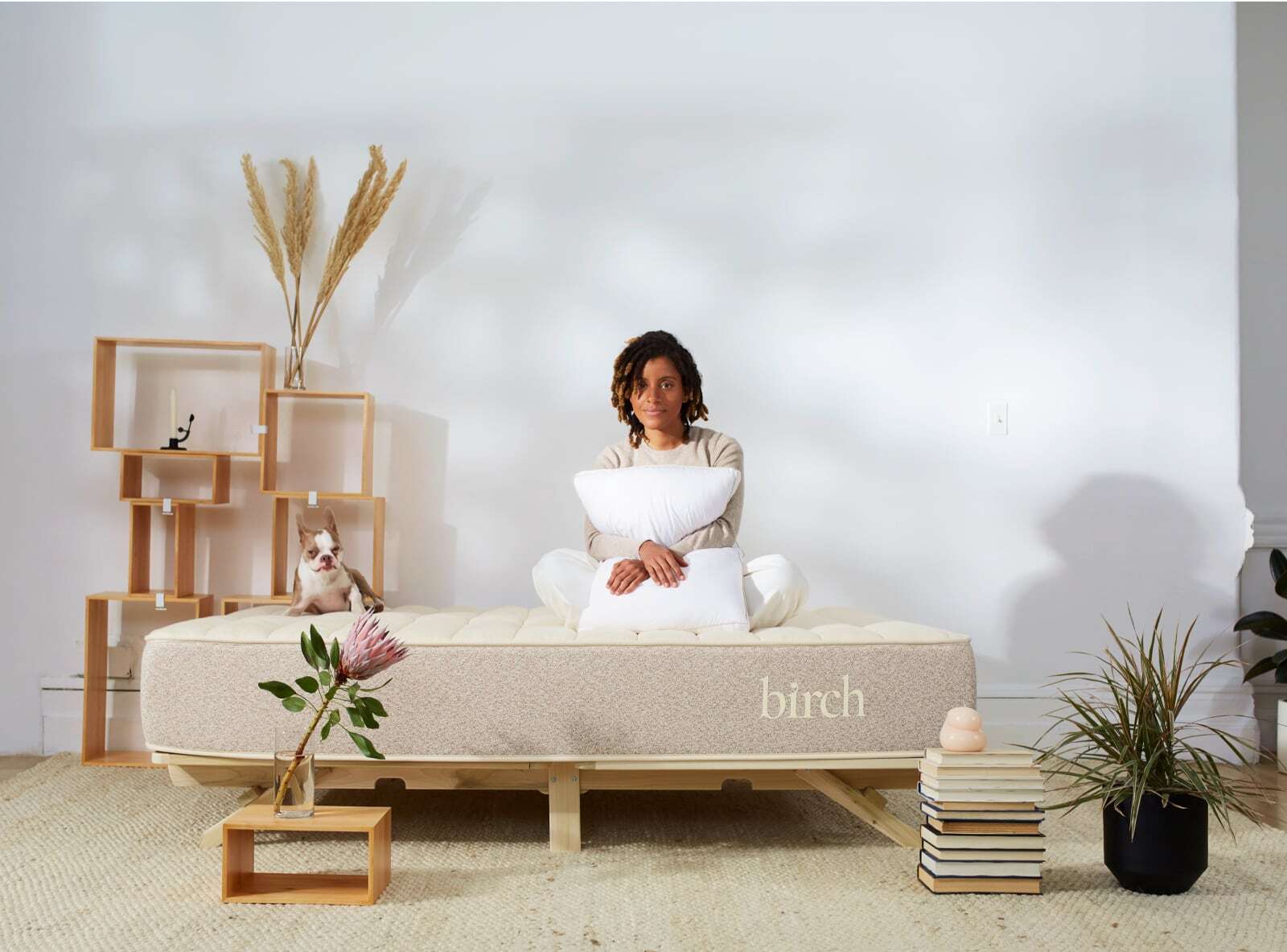 A person dressed in light-colored attire sits cross-legged on a beige Birch Natural Mattress, cradling a white pillow. They are situated in a minimalist room featuring wooden shelves, books, and a plant. A small dog is seated beside them on the mattress, gazing towards the camera.