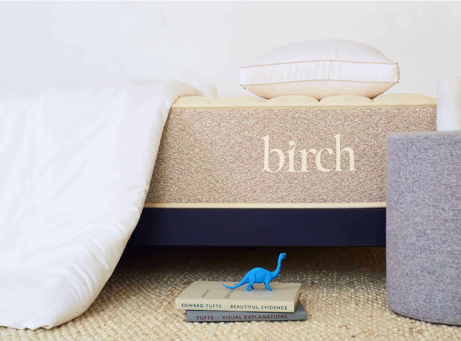 A neatly made bed with white bedding and pillows is seen in the background, with a Birch Natural Mattress visible on the side. In the foreground, a blue toy dinosaur stands on two books atop a textured, light-colored rug. A gray cylindrical stool is also present.