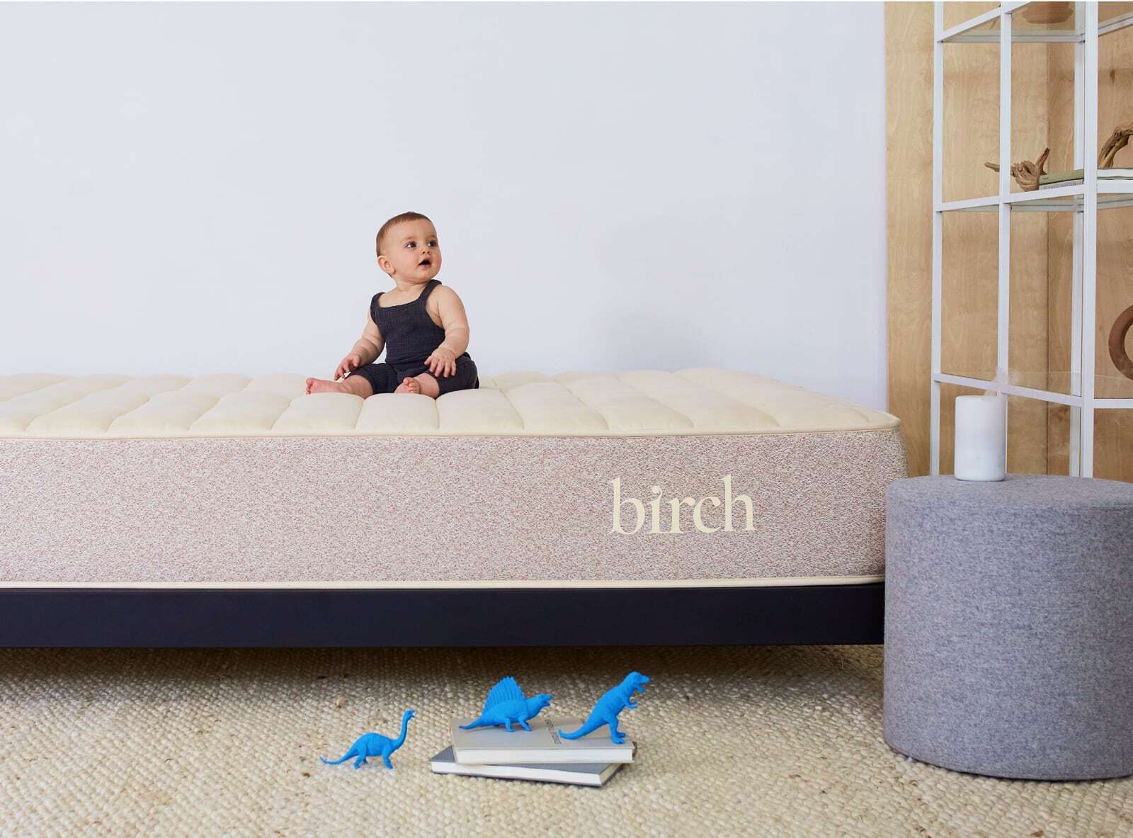 A baby wearing a dark tank top is seated on a Beige Birch Natural Mattress. In front of the mattress, blue toy dinosaurs and a children's book are placed on a woven rug. A gray cylindrical ottoman and a wooden shelf with decorative items can be seen in the background.