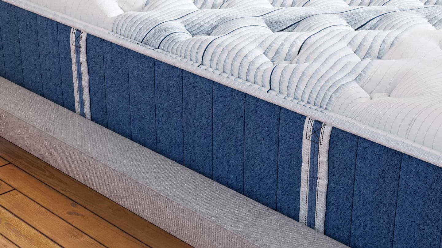 A close-up view of the Elite Hybrid mattress, showcasing its tufted white top and blue fabric sides with vertical stitched handles for easy maneuvering. The mattress rests on a grey bed frame with wooden flooring beneath.