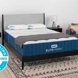 A neatly made bed featuring the Elite Hybrid mattress and pillows in elegant blue and white is centered in a modern, minimalist bedroom. Two small black nightstands with lamps flank the bed, while a gray armchair draped with a blanket sits in the background. A "2023 Good Housekeeping Best Bedding Awards" badge highlights its elite quality.