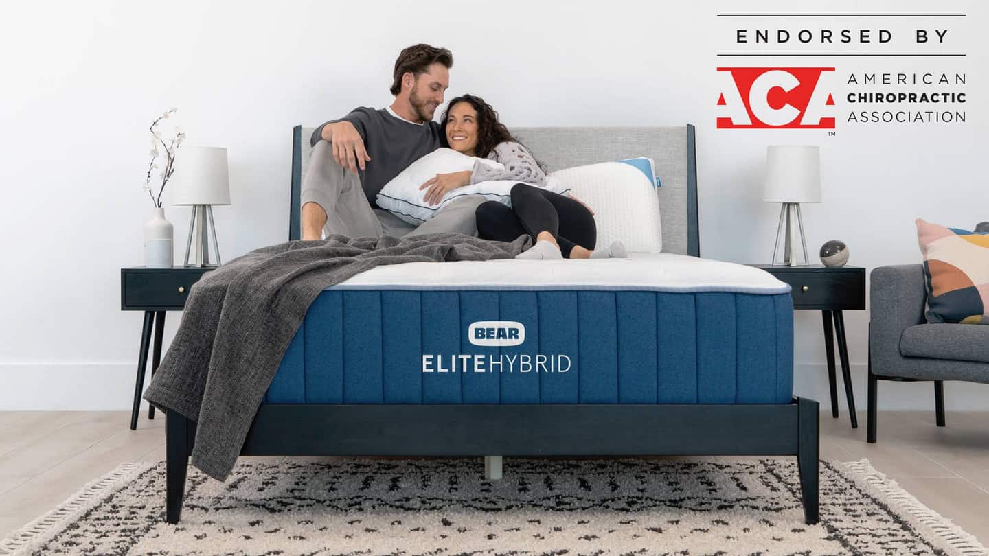A man and woman sit comfortably on a bed with a blue Elite Hybrid mattress. The room, showcasing a modern design with nightstands, lamps, and minimalist decor, emanates tranquility. The image displays the logo of the American Chiropractic Association, signifying their endorsement.