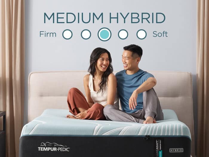 A couple sits on a Tempur-Pedic TEMPUR-LuxeAdapt® 13" Medium Hybrid Mattress in a bright room, smiling and looking at each other. The mattress, known for its medium firmness, features a firmness scale indicating medium support. Both individuals are casually dressed and appear relaxed on their comfy Tempur-Pedic hybrid mattress.