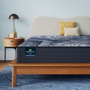 A modern bedroom featuring a Serta Perfect Sleeper X Hybrid Mattress: Kendall Plush on a wooden bed frame. The bed is adorned with textured gray bedding and yellow throw pillows. A matching wooden nightstand supports a black and gold lamp, while a blue rug lies beneath the bed.