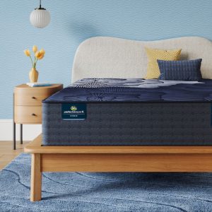 A modern bedroom featuring a bed with a Serta Perfect Sleeper X Mattress: Prescott Medium Firm mattress in dark blue and patterned bedding. The bed has a light-colored cushioned headboard and a wooden frame. A wooden nightstand with a white lamp and a vase with yellow flowers is next to the bed, all set on a blue rug.
