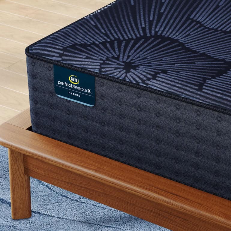 A close-up view of a Serta Perfect Sleeper X Prescott Medium Firm mattress placed on a wooden bed frame with a light blue, textured carpet underneath. The mattress features a dark blue cover with a geometric pattern.
