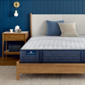 A stylish bedroom features a wooden bed frame with a cushioned headboard, neatly made with blue and beige pillows. A dark blue Serta iComfortPro Mattress: Ambrose Firm lies on the bed. A wooden nightstand with a lamp, books, and decorations is beside the bed.