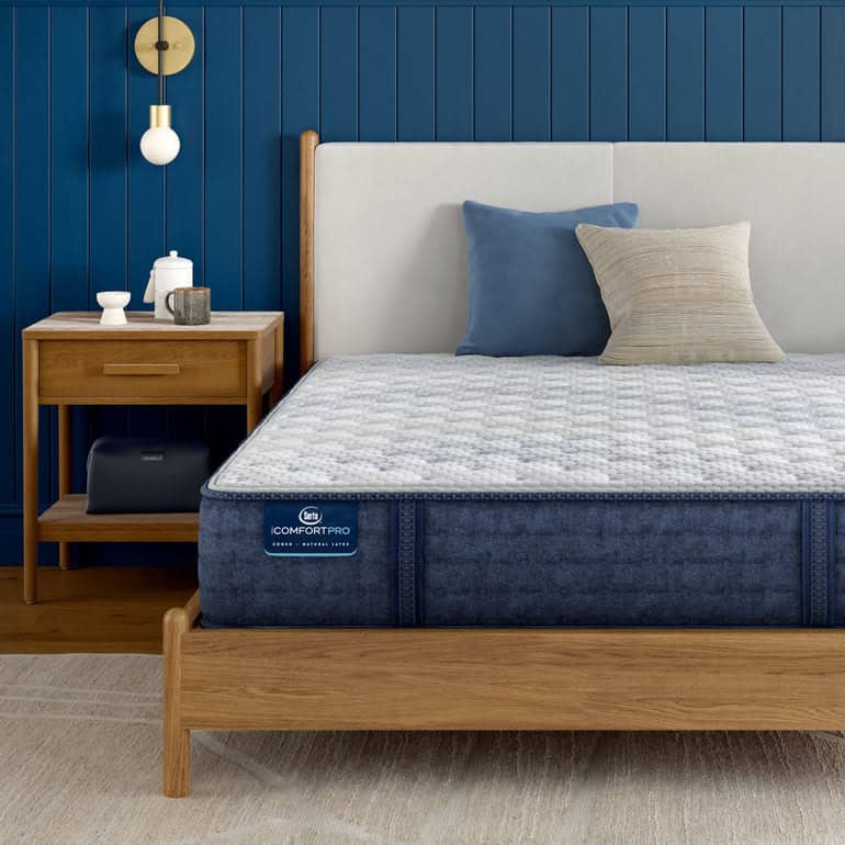 A stylish bedroom features a wooden bed frame with a cushioned headboard, neatly made with blue and beige pillows. A dark blue Serta iComfortPro Mattress: Ambrose Firm lies on the bed. A wooden nightstand with a lamp, books, and decorations is beside the bed.
