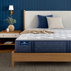 A neatly made bed features a blue and white Serta iComfortPro Mattress: Ambrose Plush with a wooden frame. The headboard is cream-colored, complemented by pillows and a throw blanket. A nightstand beside the bed holds a vase with foliage, while the wall boasts a dark blue panel design.