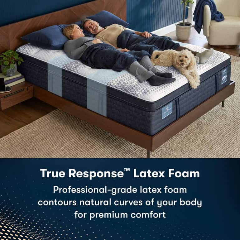 A couple is lying in bed under a blanket with a dog resting near their feet. The Serta iComfortPro Mattress: Ambrose Firm features a label indicating "True Response™ Latex Foam", promoting professional-grade latex foam that contours to the body's curves for premium comfort.