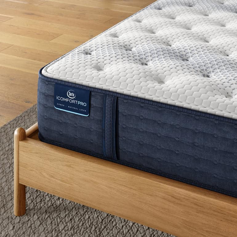 Close-up of the Serta iComfortPro Mattress: Ambrose Plush on a wooden bed frame. The mattress features a textured white top with a hexagonal pattern and a dark blue side panel showcasing the iComfort Pro logo. The background includes a light wooden floor and part of a grey rug, highlighting the plush comfort of the Ambrose model.