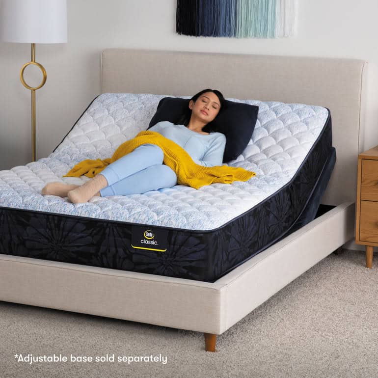 A woman relaxes on an adjustable bed with a blue-patterned Serta Classic mattress. She is lying on her back, using a black pillow, and is covered with a yellow blanket. The bed is placed in a modern, minimalist bedroom with light-colored walls and carpet, evoking a classic sense of comfort.
