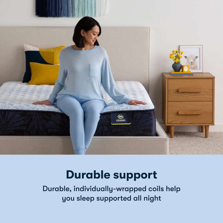 A woman in blue loungewear sits on a bed adorned with a vibrant pillow arrangement. Text on the image reads, "Durable support. The Serta Classic Mattress: Brecon Firm with individually-wrapped coils helps you sleep supported all night." Beside the bed is a wooden nightstand decorated with flowers and a clock.