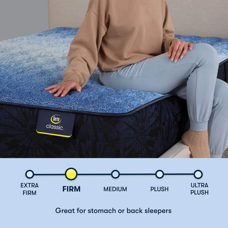 A person wearing a beige sweatshirt and light blue pants sits on the edge of a bed with a Serta Classic: Brecon Firm mattress, as indicated by the label, which is suitable for stomach or back sleepers according to the firmness scale below.