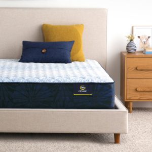 A neatly made bed with a beige upholstered frame, featuring a Serta Classic Mattress in light blue and navy, is positioned next to a wooden nightstand. The bed includes two pillows: one mustard yellow and one navy. The nightstand holds a small potted plant and framed picture.