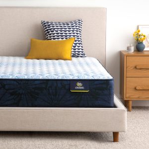 A neatly made bed featuring a Serta Classic Mattress Dorian Firm with a blue and black pattern and a white, quilted top cover. On the bed, there are two pillows: one with a blue and white wave pattern and another smaller mustard yellow pillow. Beside the bed stands a wooden nightstand adorned with flowers and holding a glass of water.