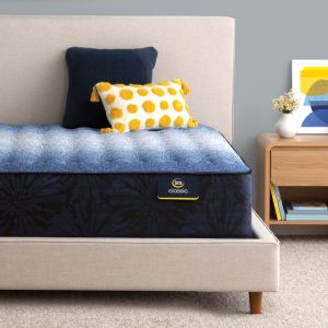 A neatly made bed with a blue and yellow polka-dot throw pillow, a navy blue pillow, and a Serta Classic Mattress: Brecon Firm sits in a minimalist bedroom. A nightstand beside the bed holds a plant, stacked books, and a framed abstract artwork.
