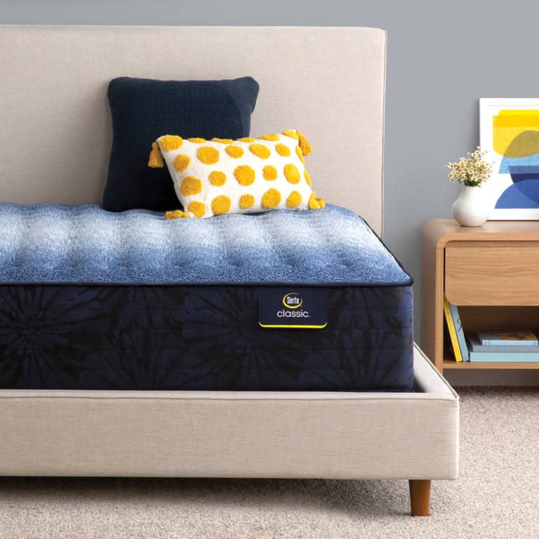 A neatly made bed with a blue and yellow polka-dot throw pillow, a navy blue pillow, and a Serta Classic Mattress: Brecon Firm sits in a minimalist bedroom. A nightstand beside the bed holds a plant, stacked books, and a framed abstract artwork.