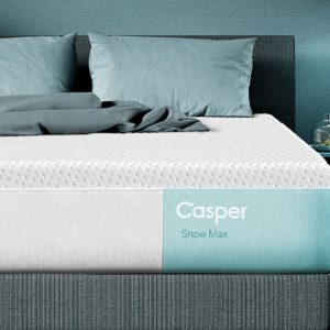A modern bedroom featuring a Casper Snow Max: Cooling Hybrid Mattress on a dark-colored bedframe adorned with teal and gray bedding. A nightstand beside the bed holds a glass lamp and a small glass orb. The room has minimalistic, cozy decor and a serene color palette, perfect for restful nights on this cooling hybrid mattress.