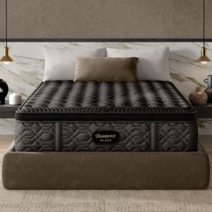 A bedroom features a neatly made bed with a Beautyrest Black® Series One 14.25" Firm Pillow Top Mattress. The bed is adorned with a beige cushion and white pillows. The modern decor includes a marble backsplash, two hanging pendant lights, and minimalist decorative items on the bedside shelf.