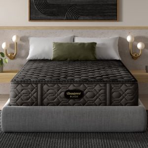 A neatly made bed with a dark, quilted Beautyrest Black® Series One 14.25" Firm Pillow Top Mattress stands at 14.25". The bed features white pillows and a green cushion against a dark headboard. Modern wall sconces with round bulbs flank the sides, while a potted plant sits on the nightstand. The room embraces a minimalist design aesthetic.