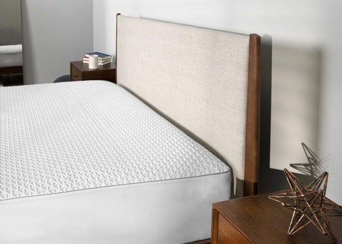 A neatly made bed featuring a Dri-Tec Mattress Protector (Copy) in a modern bedroom. The bed has a light-colored upholstered headboard with wooden trim. There are wooden nightstands on either side, one holding a stack of books, a coffee mug, and a geometric metal sculpture.