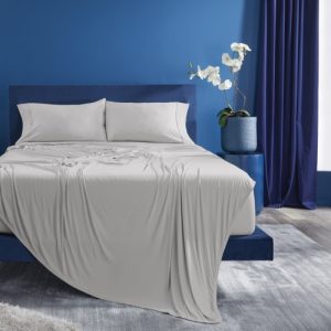 A neatly made bed with a light gray Hyper-Cotton Sheet Set (Copy) stands in a sleek, modern bedroom with blue walls and dark blue curtains. A potted white orchid sits on a bedside table, and a matching blue lamp is on the other side. The floor features a light-colored rug.
