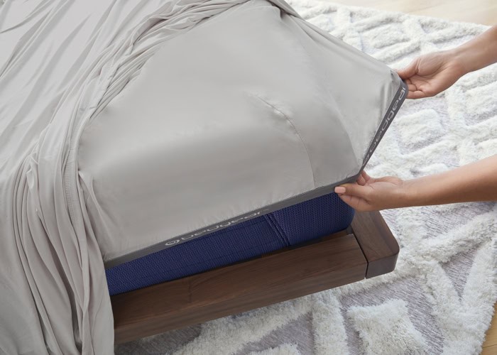 A person is adjusting a fitted Hyper-Cotton Sheet Set (Copy) on the corner of a bed. The sheet is light gray, and the bed has a wooden frame. Underneath the sheet, a blue mattress is visible. The bed is on a soft, textured, light-colored rug.