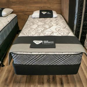 A neatly arranged display of two twin-sized beds with decorative bedding, labeled "Spring Air Back Supporter." The beds are set on a wooden floor with a rustic wooden backdrop. A stack of similar mattresses is visible on the right side.