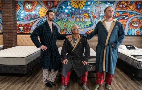 Three people wearing robes stand and sit on mattresses in a room with colorful, mural-covered walls depicting abstract cityscapes and nature.