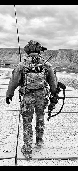 A soldier in camouflage gear holds a rifle and wears a backpack adorned with symbols. He stands on a patterned surface, facing rocky terrain under a cloudy sky.
