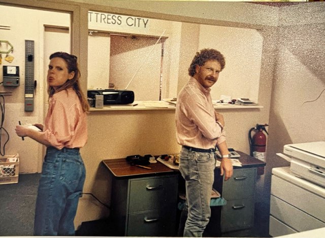 A man and woman in a vintage office. The woman stands near an old phone, and the man, with curly hair, is near a desk with a copier. Both wear casual outfits. A wall plaque reads "MATTRESS CITY." Various office items are in the background.