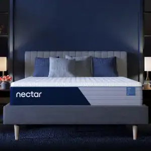 A neatly made bed with a grey tufted headboard and a grey Nectar Ultra Memory Foam Mattress is flanked by two wooden nightstands, each holding a white lamp. The room features dark blue walls and large windows.