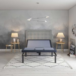 A modern bedroom showcases a gray Bed Tech 3000 Adjustable Base on a geometric rug, flanked by two wooden side tables with lamps. The walls feature a cloudy gray paint effect, and a contemporary pendant light hangs from the ceiling.