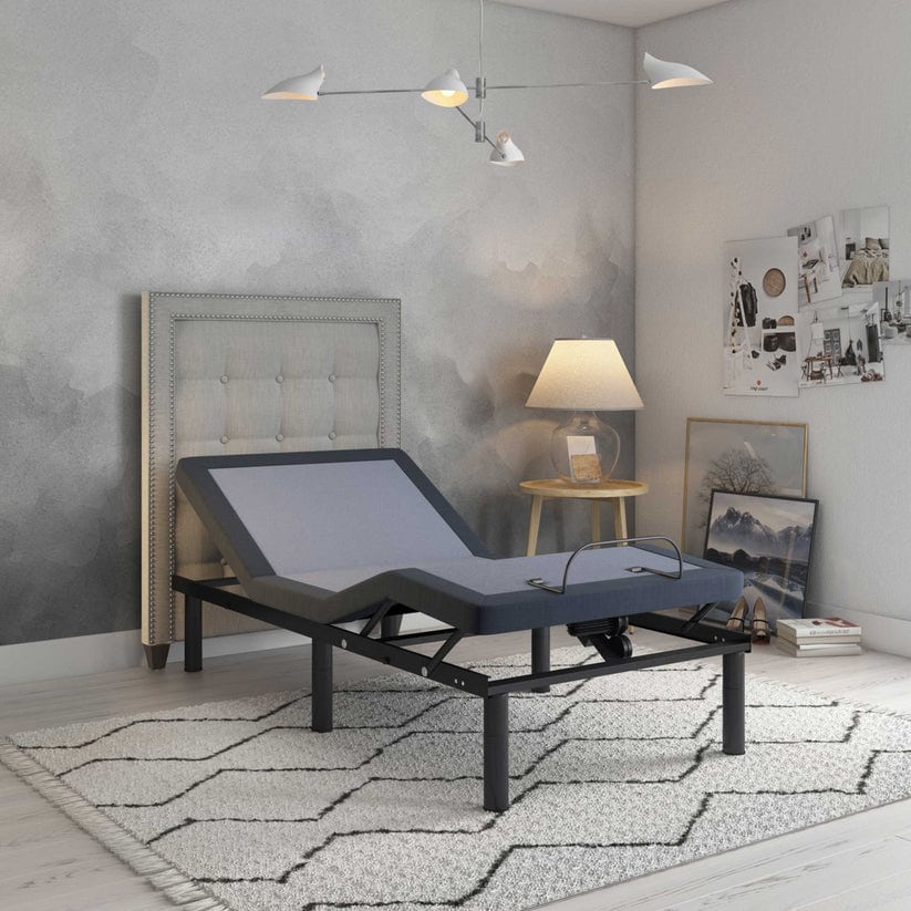 A modern bedroom highlights the Bed Tech 3000 Adjustable Base with a gray upholstered headboard, geometric rug, side table with lamp, casually placed artwork against the walls, and a minimalist ceiling light fixture hanging above.