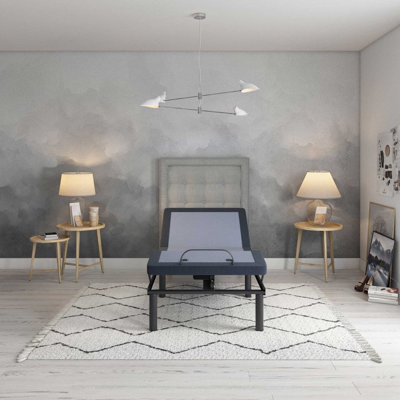 A modern bedroom features the gray Bed Tech 3000 Adjustable Base on a white geometric rug. Two wooden side tables with lamps flank the bed. Abstract art and photos adorn the walls, while a minimalist chandelier hangs from the ceiling.