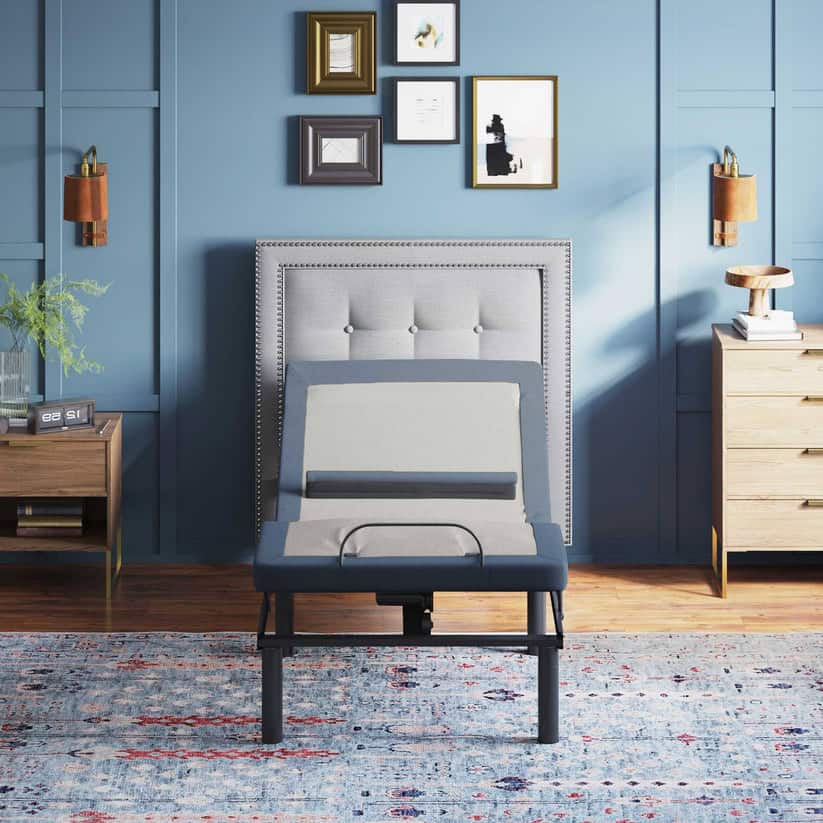 A modern bedroom has a blue accent wall and a patterned rug. The Glideaway King Glide-A-Matic bed frame features a grey upholstered headboard with an adjustable mattress raised at both ends. Two wooden nightstands hold lamps, and framed art is displayed above the bed.