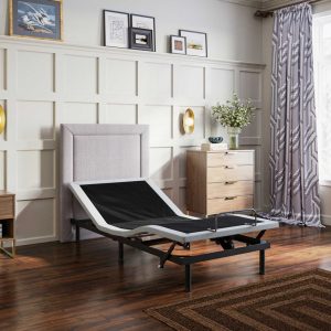 The modern bedroom features a Bed Tech X4 Adjustable Base as its centerpiece, with a wooden floor, beige upholstered headboard, dresser holding a vase of flowers, patterned curtains, and framed artwork on the wall.