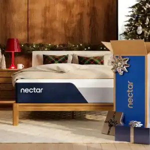 A bedroom features a Copper by Spring Air Hybrid Quilted mattress on a wooden bed frame. Decorated for Christmas, the room has a quilted throw, festive pillows, gifts like a large blue box with silver bow under the tree, and a wooden nightstand with a red lamp.