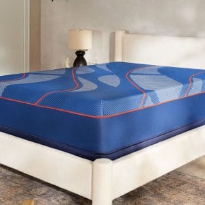 A modern bedroom showcases a white upholstered bed frame with a Sealy Posturepedic® Elite Hybrid Brenham II 14" Firm Mattress featuring wavy patterns and red trim, set against a neutral wall with a lamp and decor on the bedside table.