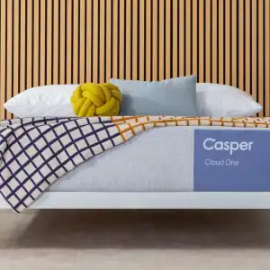 A neatly made bed features a Casper "Cloud One" Mattress with white and blue bedding, a geometric blanket, plush pillows, a yellow knot accent pillow, all set before a modern wood slat wall.