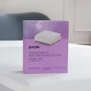 A box of Purple SoftStretch Sheets rests on a bed with a white mattress cover. The purple box shows folded bedding and notes that the sheets fit standard or queen size mattresses.