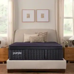 A neatly made bed features the thick, dark-colored PurplePlus Mattress (Copy) in a cozy bedroom with beige walls, wood nightstands, and large windows dressed with white curtains. Two framed pictures are displayed above the upholstered headboard.