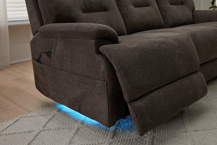 A close-up of a Zofa Umber Power Sleep Sofa with one footrest extended and blue LED lights glowing underneath, set on a light-textured rug in a living room with wooden flooring.