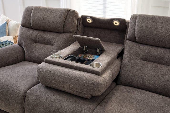 A Zofa Umber Power Sleep Sofa in gray features a center console with an open compartment holding cards, cup holders, and speakers in the headrest. Sunlight shines through a window behind the sofa.