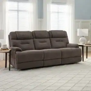 A Zofa Umber Power Sleep Sofa rests on a light patterned rug in a sunlit living room, between two round side tables topped with white lamps and decor. Large windows with sheer curtains and a tall potted plant are behind.