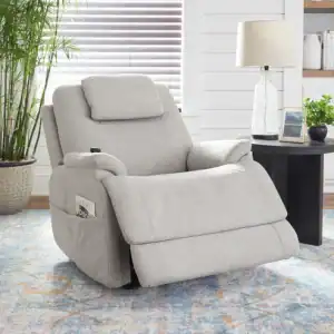 A light gray Zecliner Model 2 Petite — Power Sleep Chair with textured fabric sits partially reclined in a living room near a round black side table with a lamp and plant, a potted bamboo, and a window with white blinds.